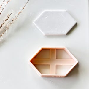 May include: Two hexagon-shaped cookie cutters. One is white, solid, and smooth. The other is light pink, with a cross-shaped interior design. The cutters are on a white surface, with a few sprigs of dried grass in the upper left corner.