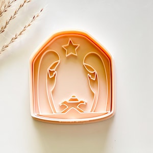 May include: A light peach-colored cookie cutter in the shape of a nativity scene. The design includes outlines of Mary, Joseph, the baby Jesus in a manger, and a star. The cutter has a rectangular shape with rounded edges.
