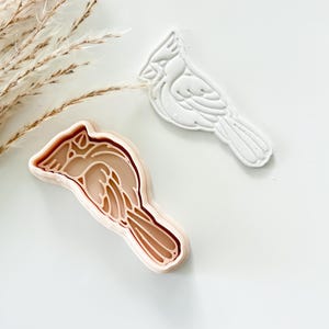 May include: A light peach-colored plastic cookie cutter shaped like a cardinal bird, featuring an internal stamp design for detailed wing and feather impressions, alongside a white molded dough cutout of the same bird shape.