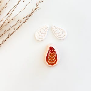 May include: Three oyster-shaped clay pieces in a studio setting. Two are white, shell-shaped, and the third is a pinkish-beige color with a reddish-brown interior. The objects are arranged on a white surface with dried grass in the upper left corner.