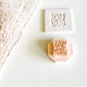 May include: A light pink, octagonal stamp with a floral design and a white, square stamp with a similar design. The stamps are on a white surface, with dried ornamental grass in the background.