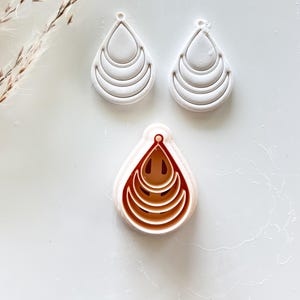 May include: Three white teardrop-shaped earring cutters with layered, curved details are displayed. A fourth cutter, in a contrasting reddish-brown, is also visible. The cutters are arranged on a white surface, suggesting a crafting or jewelry-making theme.
