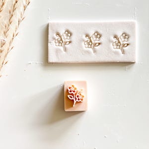 May include: A rectangular stamp and a block with three floral designs. The stamp is a light peach color with a white floral design. The block is a light pink color with three floral designs.