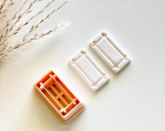 Peut inclure: Un emporte-pièce rectangulaire orange clair et rose avec un motif détaillé, à côté de deux biscuits rectangulaires. L'emporte-pièce a un cadre et des supports intérieurs. Les biscuits sont de couleur claire et ont un design de cadre similaire.