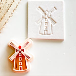 May include: A windmill-shaped cookie cutter and its impression on a white surface. The cutter is orange and white, with a detailed design of a windmill. The impression is white, showing the windmill's outline. The image is on a white background.