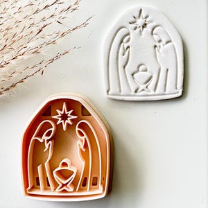 May include: A peach-colored plastic cookie cutter featuring a nativity scene with two figures standing over a manger beneath a star, accompanied by a white stamped impression of the same design on a flat surface.