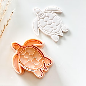 Peut inclure: Deux emporte-pièces en forme de tortue. L'un est blanc, montrant les détails de la carapace de la tortue. L'autre est de couleur pêche, avec un intérieur blanc, représentant également une tortue. Pour la pâtisserie ou l'artisanat.