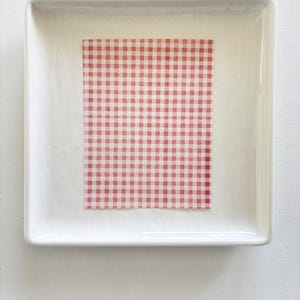 May include: A white rectangular ceramic dish with a red and white gingham patterned paper inside. The dish has slightly rounded corners and a glossy finish. The paper is centered within the dish.