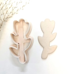 May include: Two white plastic cookie cutters shaped like stylized plant leaves. The cutters are designed to create cookies with a detailed leaf pattern.