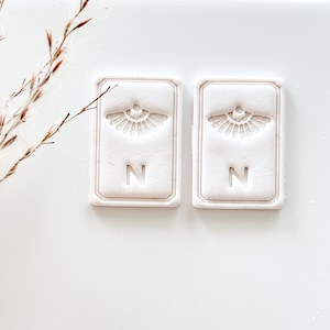 May include: Two rectangular, off-white decorative tiles with a raised fan-like design and the letter "N". The tiles have a textured surface and a raised border. The background is white.