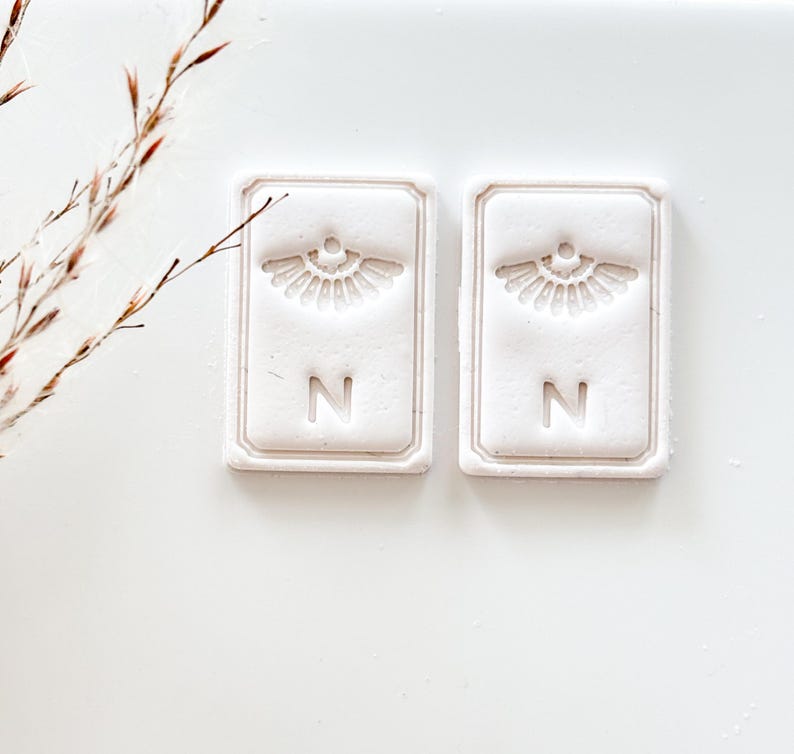 May include: Two rectangular, off-white decorative tiles with a raised fan-like design and the letter "N". The tiles have a textured surface and a raised border. The background is white.