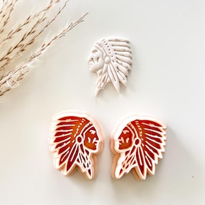 May include: Three cookie cutters in the shape of a Native American chief's profile. Two cutters are red and white, and one is white. The cutters are on a white surface with a sprig of dried grass.