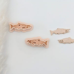 May include: A collection of salmon-shaped cookie cutters in varying sizes. The cutters are a light brown color with detailed outlines, including gills and fins. The design is layered, creating a 3D effect. Ideal for baking or crafting.