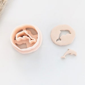 May include: Three pink plastic cookie cutters shaped like dolphins. The largest cutter is a single dolphin, the medium cutter is a single dolphin, and the smallest cutter is a double dolphin.