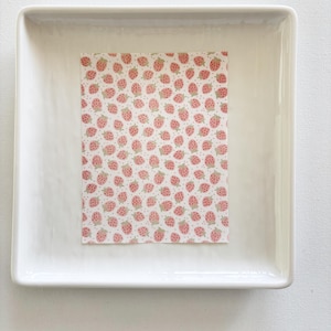 May include: A white rectangular ceramic tray contains a rectangular piece of paper. The paper is patterned with small pink strawberries and green stems on a white background. The tray is on a white surface.