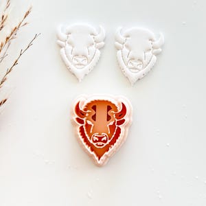 May include: Three bison-shaped cookie cutters. Two are white, and one is a layered design with orange and red details. The cutters are arranged on a white surface, showcasing the bison's head and horns.