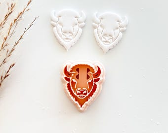 May include: Three bison-shaped cookie cutters. Two are white, and one is a layered design with orange and red details. The cutters are arranged on a white surface, showcasing the bison's head and horns.