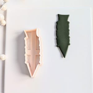 May include: A close-up of a cookie cutter in the shape of a pencil. The cutter is made of white plastic and is shown next to a green fondant cutout of the same shape.