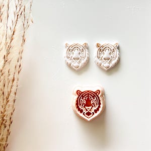 May include: Three tiger head-shaped cookie cutters. Two are light beige, and one is red and beige. The tiger faces have detailed facial features. The cutters are on a white surface.