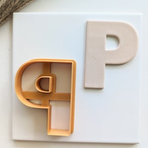 May include: A close-up of two letter P cookie cutters. One is a solid beige color and the other is a bright orange plastic. Both are on a white background.