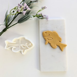 May include: A white plastic cookie cutter shaped like a fish, and a gold-colored cookie in the shape of a fish on a white marble surface.