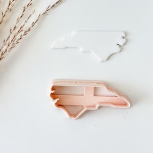 May include: A white North Carolina state-shaped cookie cutter and a matching light peach-colored plastic cutter frame. The white piece is a solid silhouette of the state, while the peach piece is the open-frame tool used for cutting the dough shape.