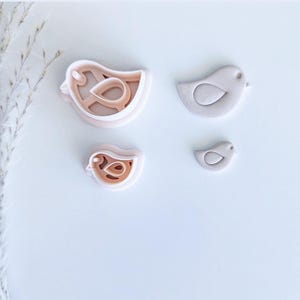 May include: Four bird-shaped cookie cutters in varying sizes. Two are layered, showing the cutting and interior design, while the other two are solid. The cutters are light pink and tan, ideal for baking themed treats.
