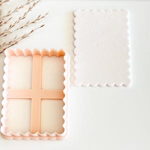 May include: A light pink cookie cutter with a scalloped edge and a cross-shaped interior, next to a matching light pink rectangular shape with a scalloped edge. The items are on a white surface.