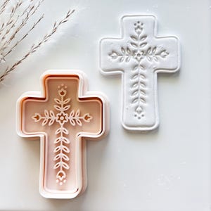 May include: Two cross-shaped cookie cutters. One is a light pink color, and the other is white. Both have a floral design in the center. The cookie cutters are on a white surface.