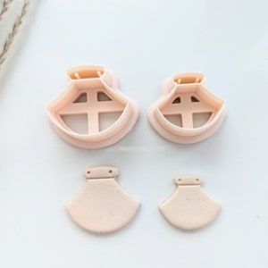 May include: Two sets of pink plastic cookie cutters in the shape of a skirt. The larger cutter has a wider skirt and the smaller cutter has a narrower skirt. Both cutters have a small hole at the top.