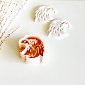 May include: An orange eagle head cookie cutter next to two white eagle head cookies. The cookie cutter is made of plastic and has a detailed design. The cookies are made of a white dough and have a similar design.