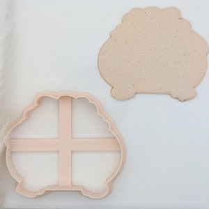 May include: A pink plastic cookie cutter in the shape of a pumpkin with a cross-hatch design inside. A plain, light brown cookie is shown next to the cutter.