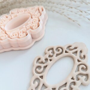 May include: Two pink plastic cookie cutters in the shape of an ornate oval frame. The cutters are on a white surface.