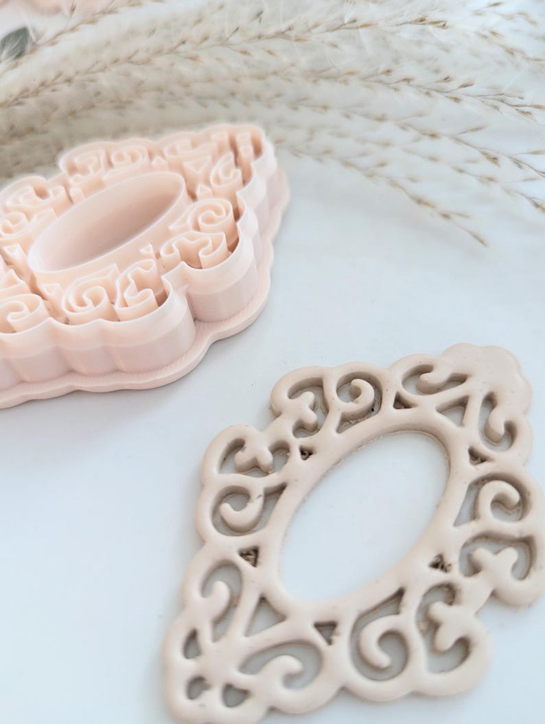 May include: Two pink plastic cookie cutters in the shape of an ornate oval frame. The cutters are on a white surface.