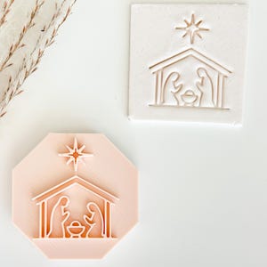 May include: Two Christmas-themed stamps. One is a light pink octagon shape, and the other is a white square. Both stamps feature a nativity scene with a star above the stable.
