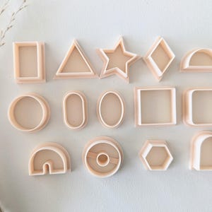 May include: A set of 15 plastic cookie cutters in various shapes, including circles, squares, rectangles, stars, triangles, and arches. The cutters are all a light beige colour.