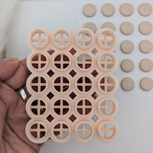 May include: A pink plastic mould with 25 round cutouts, each with a cross-shaped design. The mould is used for making cookies or other treats.