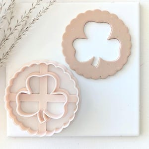 May include: Two plastic cookie cutters in the shape of a shamrock. The cutter on the left is solid and the cutter on the right is a cutout.