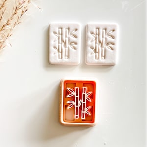 May include: A bamboo-themed cookie cutter with two pressed cookies and a cutter. The cutter is orange with a white bamboo design. The cookies are rectangular and white, with a bamboo design pressed into them.