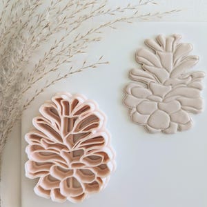 May include: Two pink plastic cookie cutters shaped like pine cones. The cutters are on a white surface.