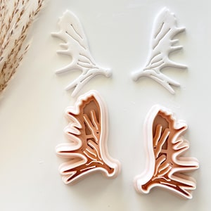 May include: Four antler-shaped cookie cutters. Two are white, and two are layered with shades of brown and pink. The cutters are arranged on a white surface, suggesting a crafting or baking theme. The design is detailed and precise.