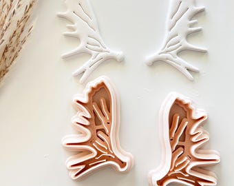May include: Four antler-shaped cookie cutters. Two are white, and two are layered with shades of brown and pink. The cutters are arranged on a white surface, suggesting a crafting or baking theme. The design is detailed and precise.