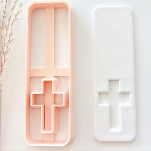 May include: A light pink plastic cookie cutter in the shape of a cross, next to a white rectangular shape with a cross imprint. The cookie cutter has a rectangular frame with a cross shape inside.