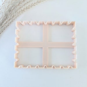 May include: A pink plastic cookie cutter shaped like a rectangle with four squares inside. The cookie cutter has a scalloped edge.