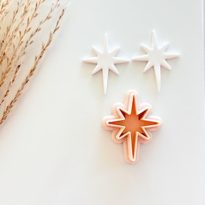 May include: Three star-shaped cookie cutters. Two are white, and one is a layered design with a light orange interior and a pinkish-orange outline. The cutters are on a white surface, with dried ornamental grass in the upper left corner.