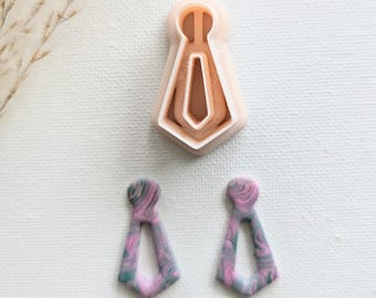 May include: A pair of pink and gray marbled tie-shaped earrings with a cutout design. The earrings are next to a plastic mold of a tie.