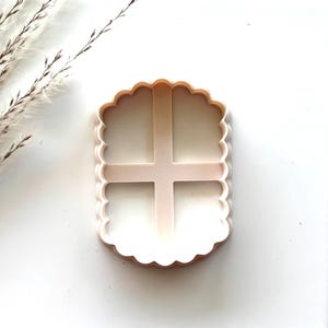 May include: A white plastic cookie cutter shaped like a rectangle with a scalloped edge and four squares inside.