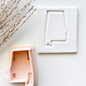 May include: A light peach-colored plastic cookie cutter shaped like the state of Alabama, positioned next to a white square piece of clay or dough that has been embossed with the same Alabama state outline shape.