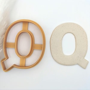 May include: A gold plastic cookie cutter in the shape of the letter Q, next to a white ceramic letter Q.