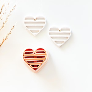 May include: Three heart-shaped objects. Two are white with horizontal lines and a small hole at the top. The third is a pink heart with a red interior and horizontal lines. The objects are on a white surface.